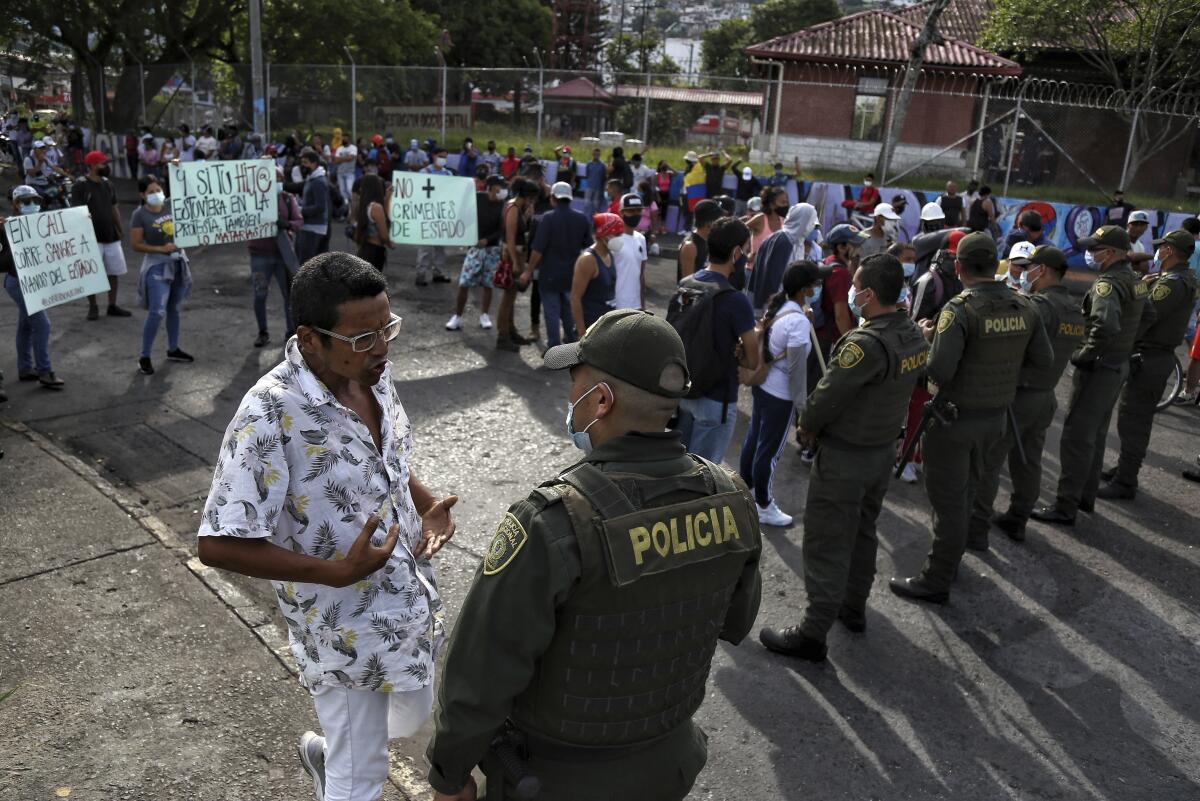 A man in a white patterned shirt and glasses stands in front of a line of police as protesters hold signs.