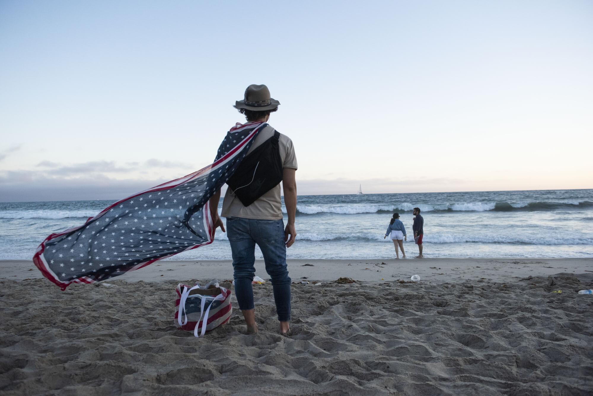 A beachgoer with a cape in U.S. flag colors stands on the sand looking out toward waves 