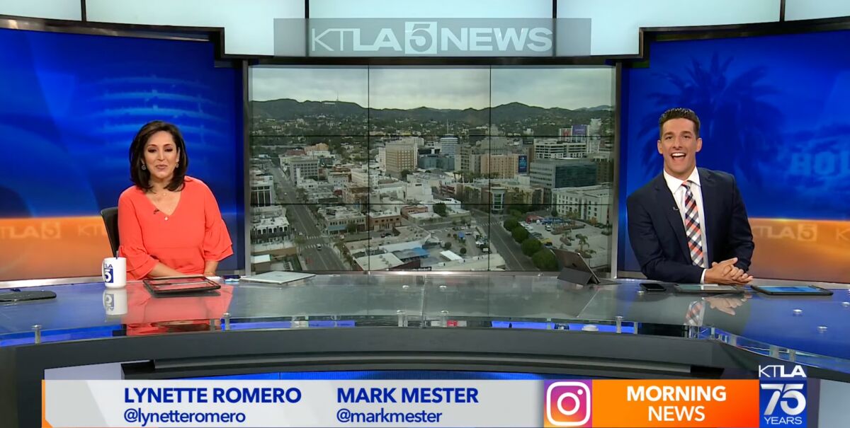 Two news anchors sit at a desk with a video backdrop behind them