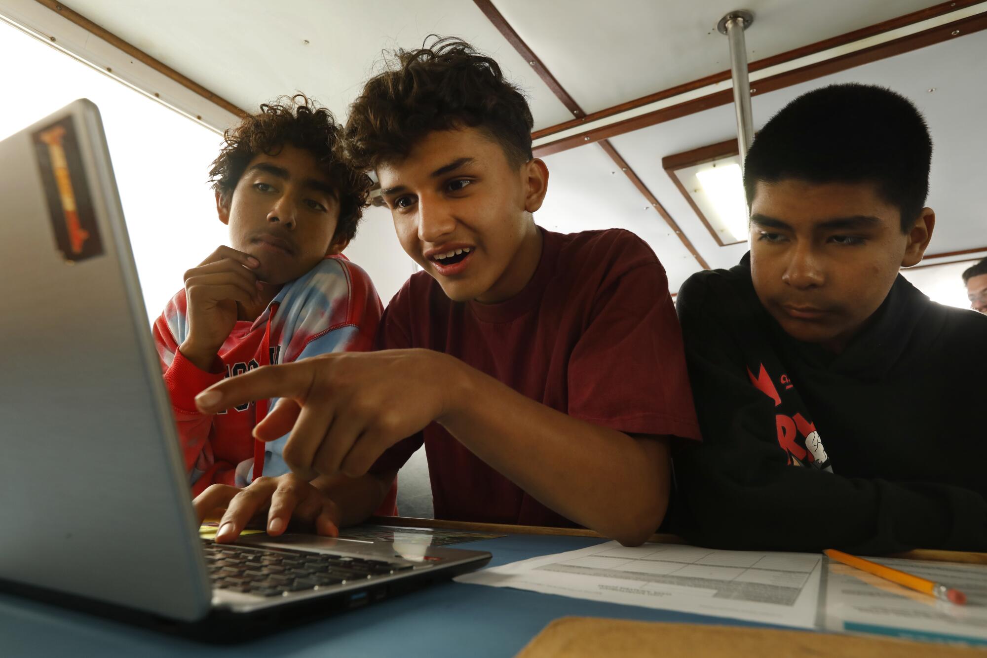 Three teens aboard a boat look at a laptop screen.