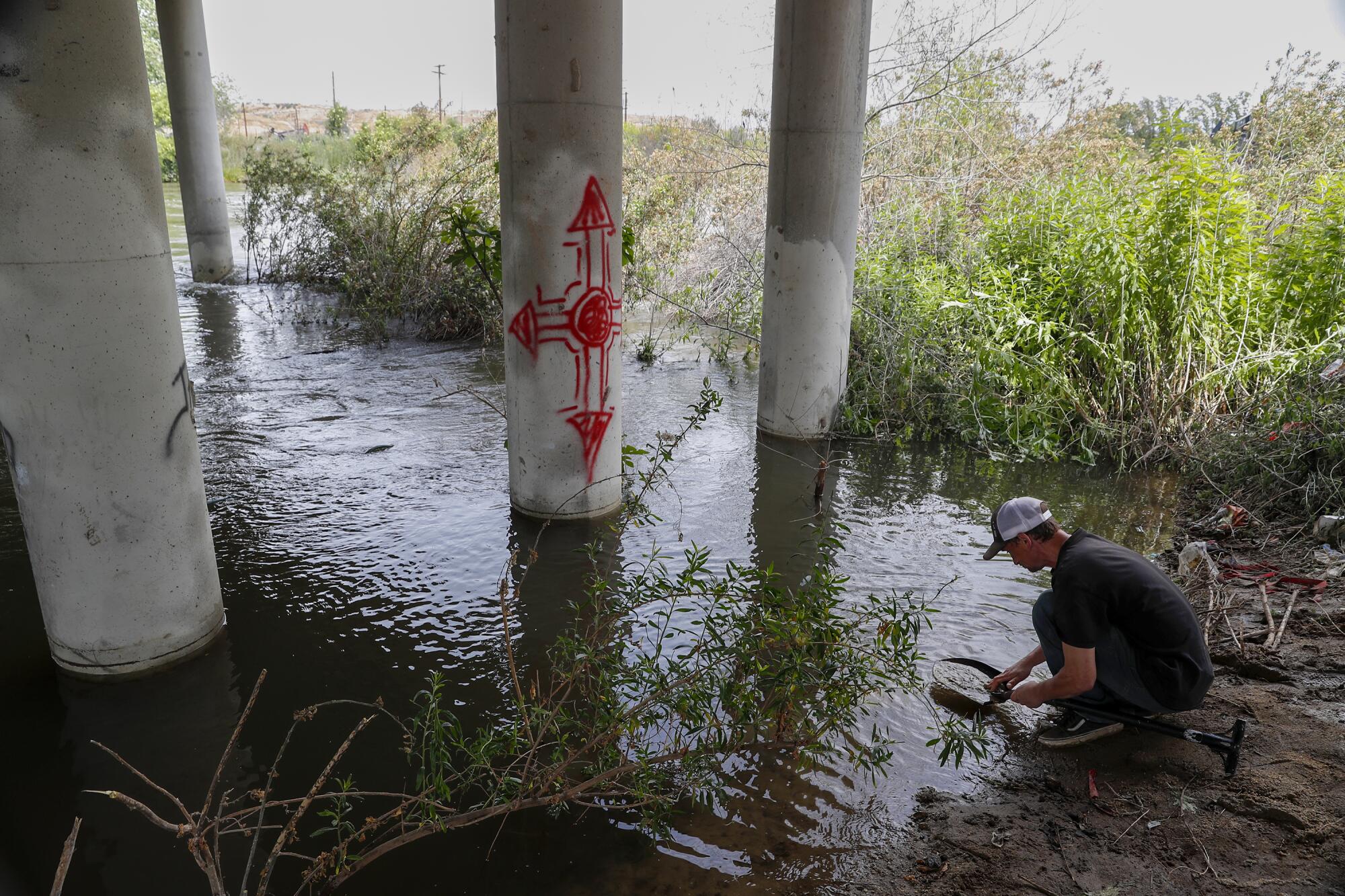 A man in a baseball cap and t-shirt pans for gold beneath a bridge.