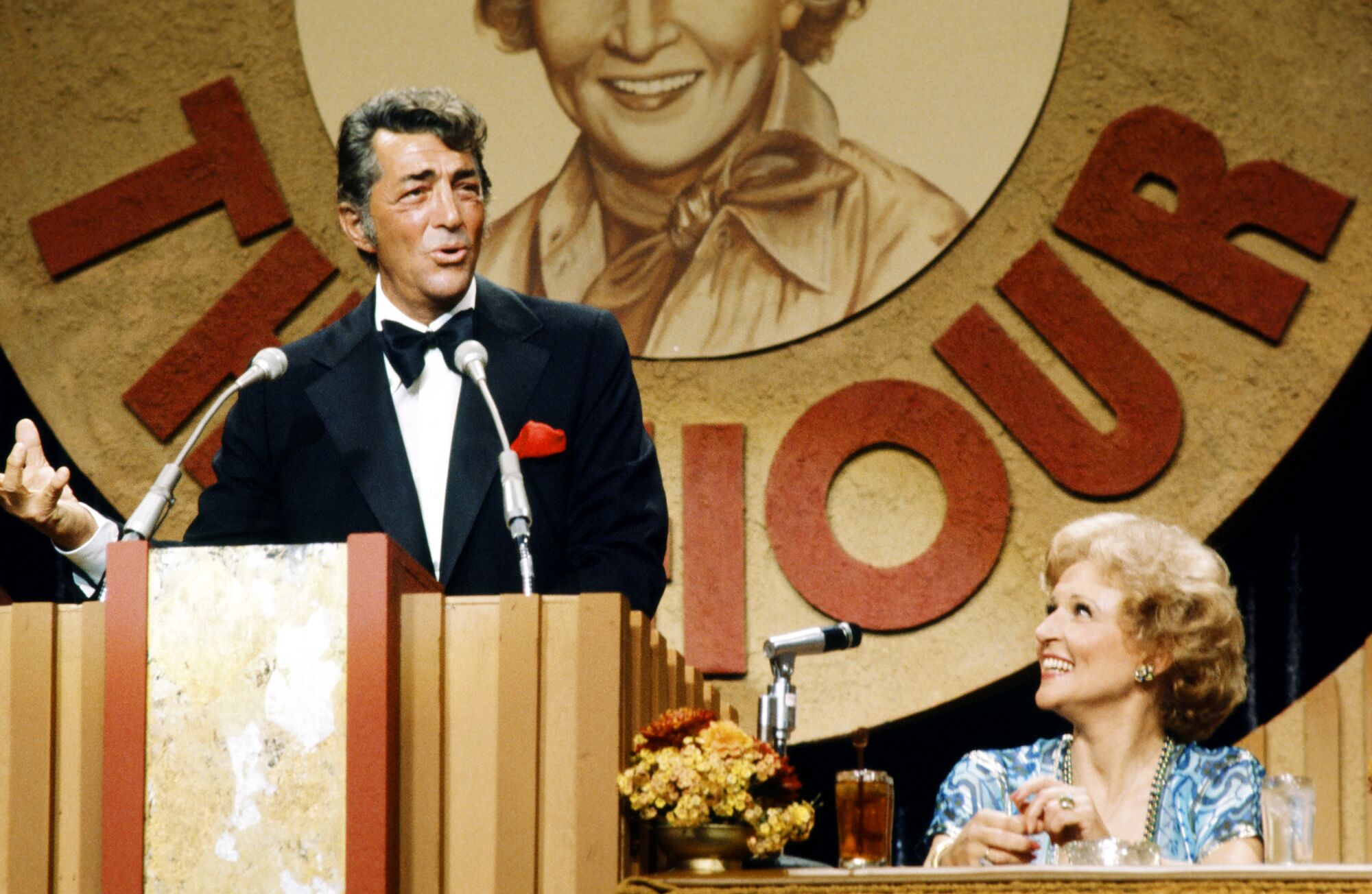 A man hosting a celebrity roast stands behind a podium as female roastee looks on from dais.