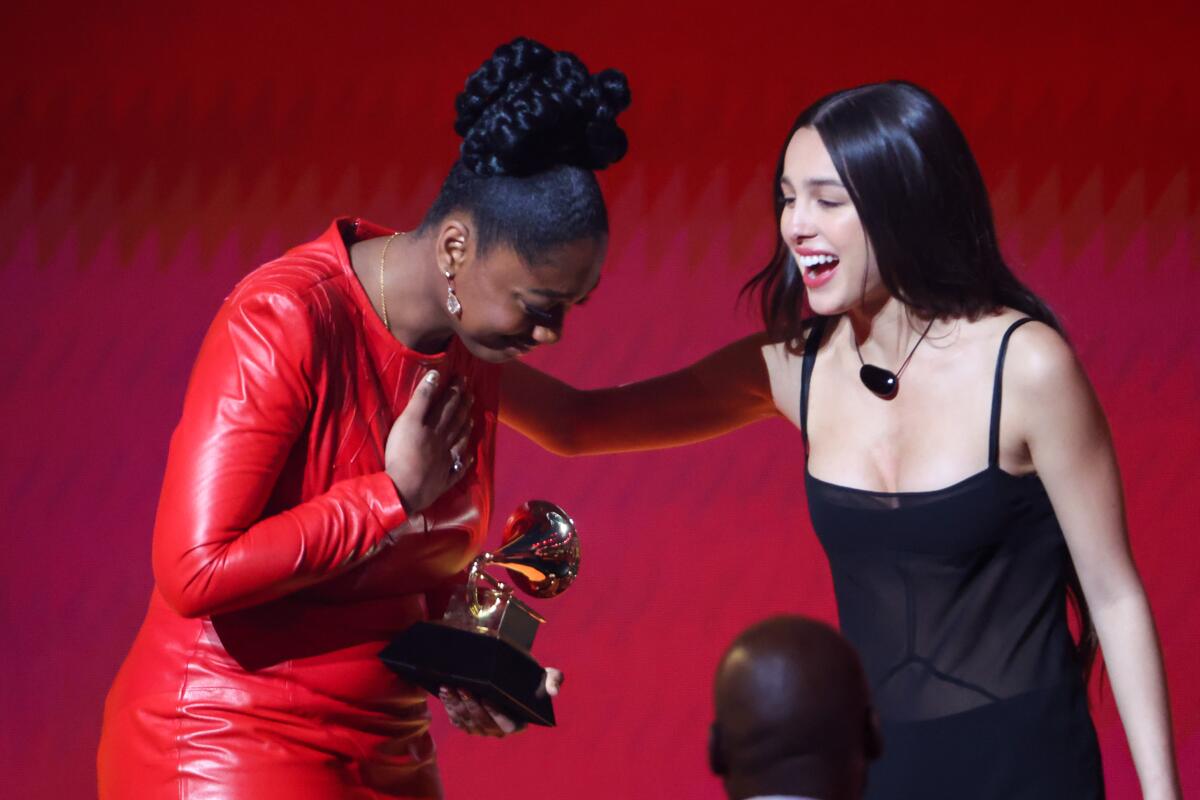 A woman in a red dress holds a Grammy while a woman in a black dress places a hand on her shoulder.