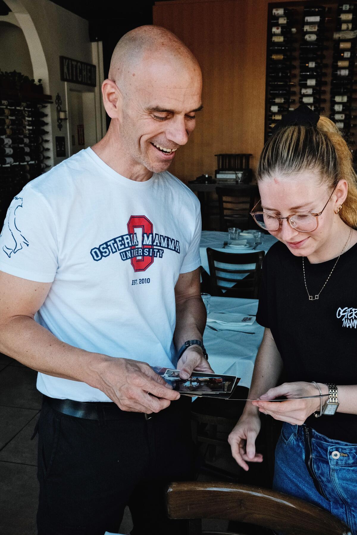 A man and a young woman stand together, smiling, looking down at photos held in their hands in a restaurant dining room.