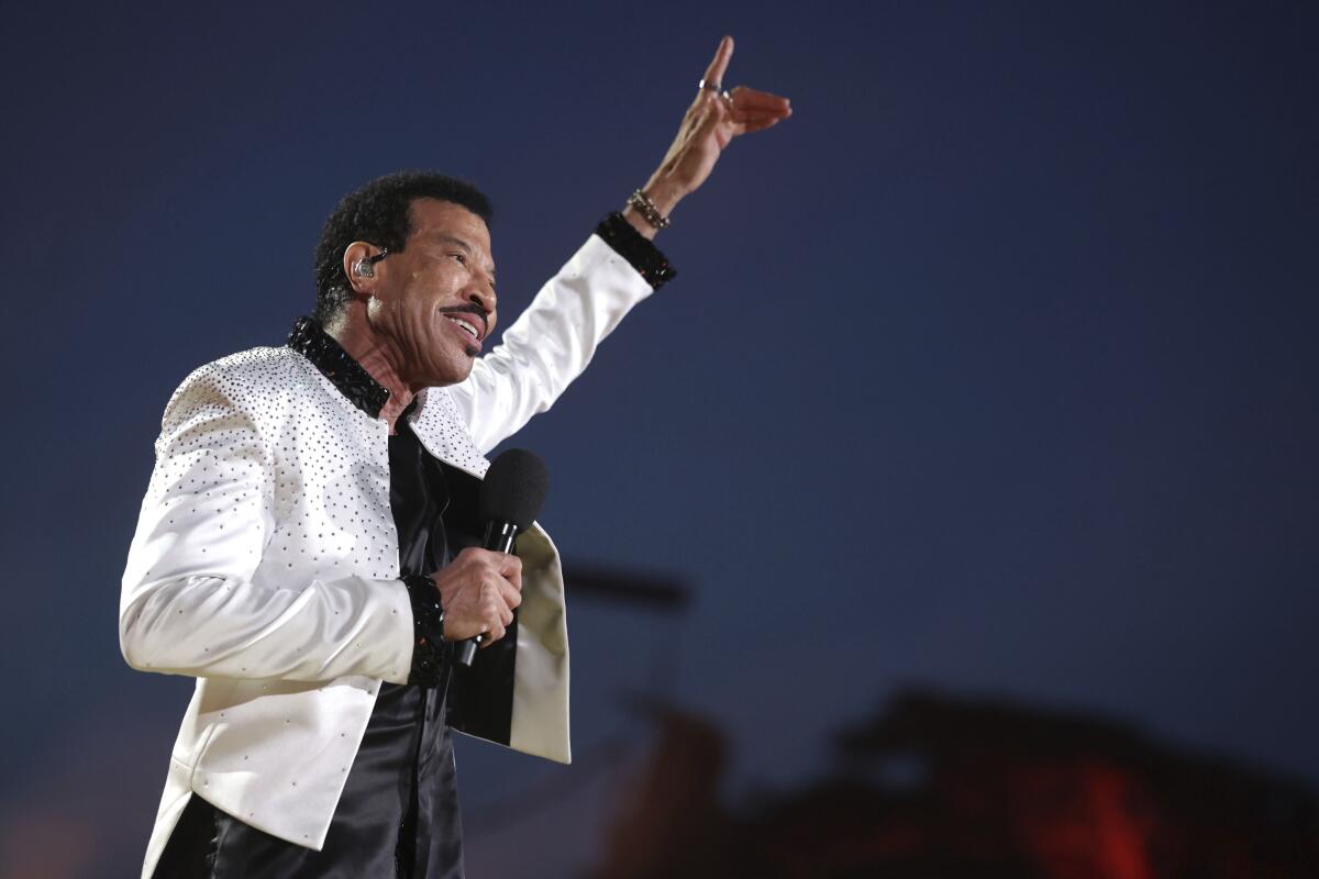 Lionel Richie holds a microphone and points toward the sky in a bedazzled white jacket and black pants