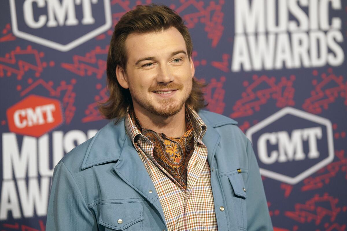 Morgan Wallen with long, brown hair posing in a blue jacket and plaid shirt