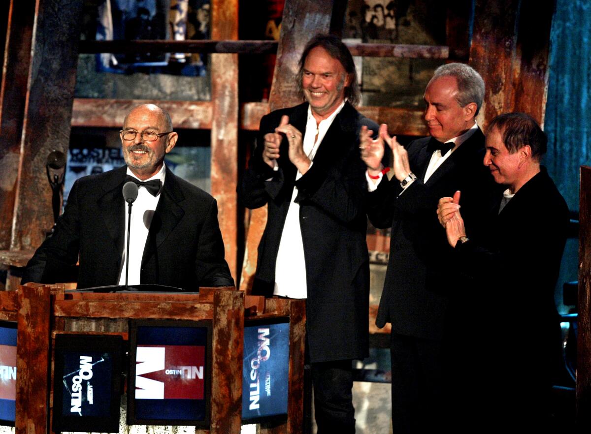 A man in a tuxedo stands in front of a podium, as three other men in tuxes stand nearby and applaud.