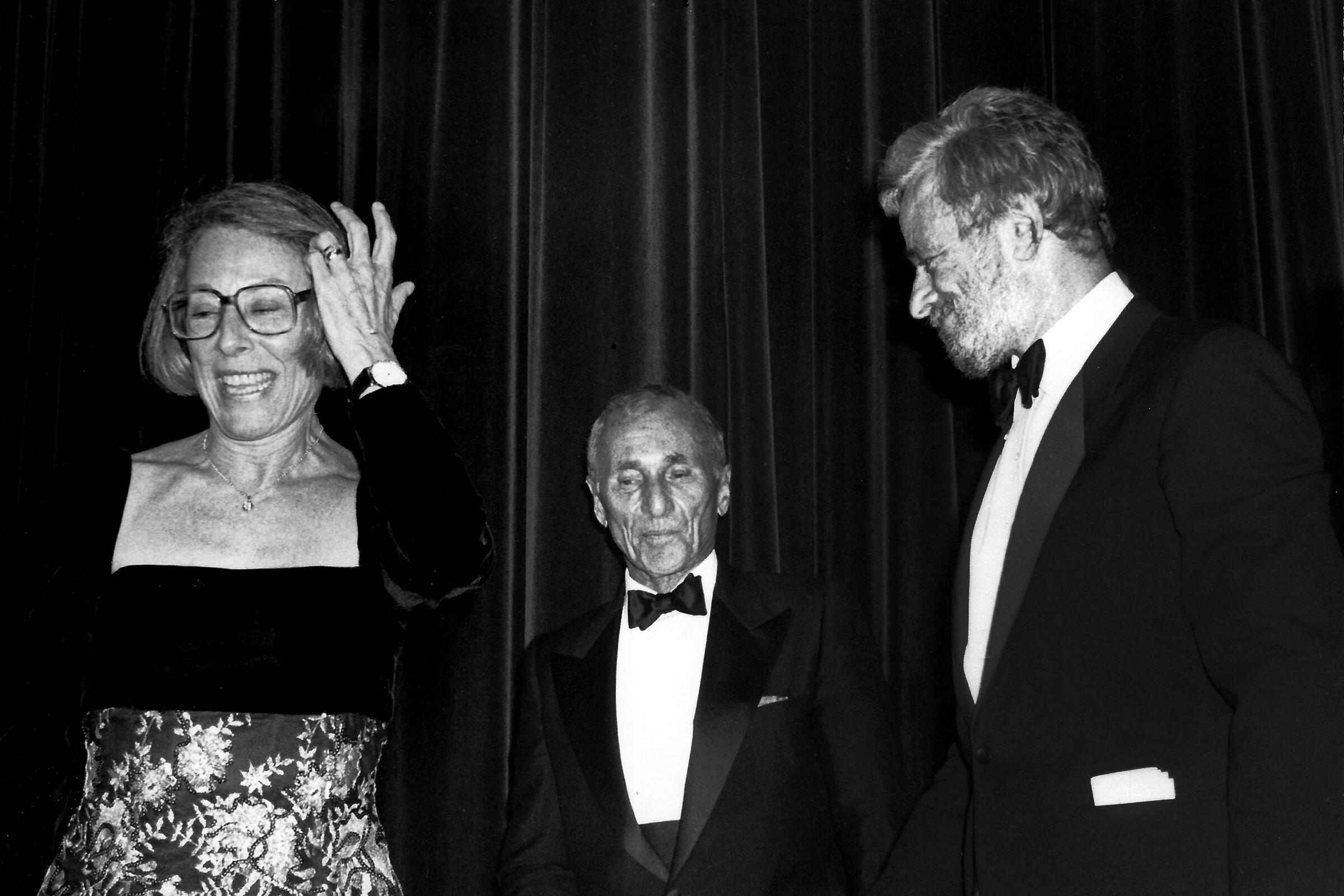 A woman adjusts her hair next to two men in tuxes.