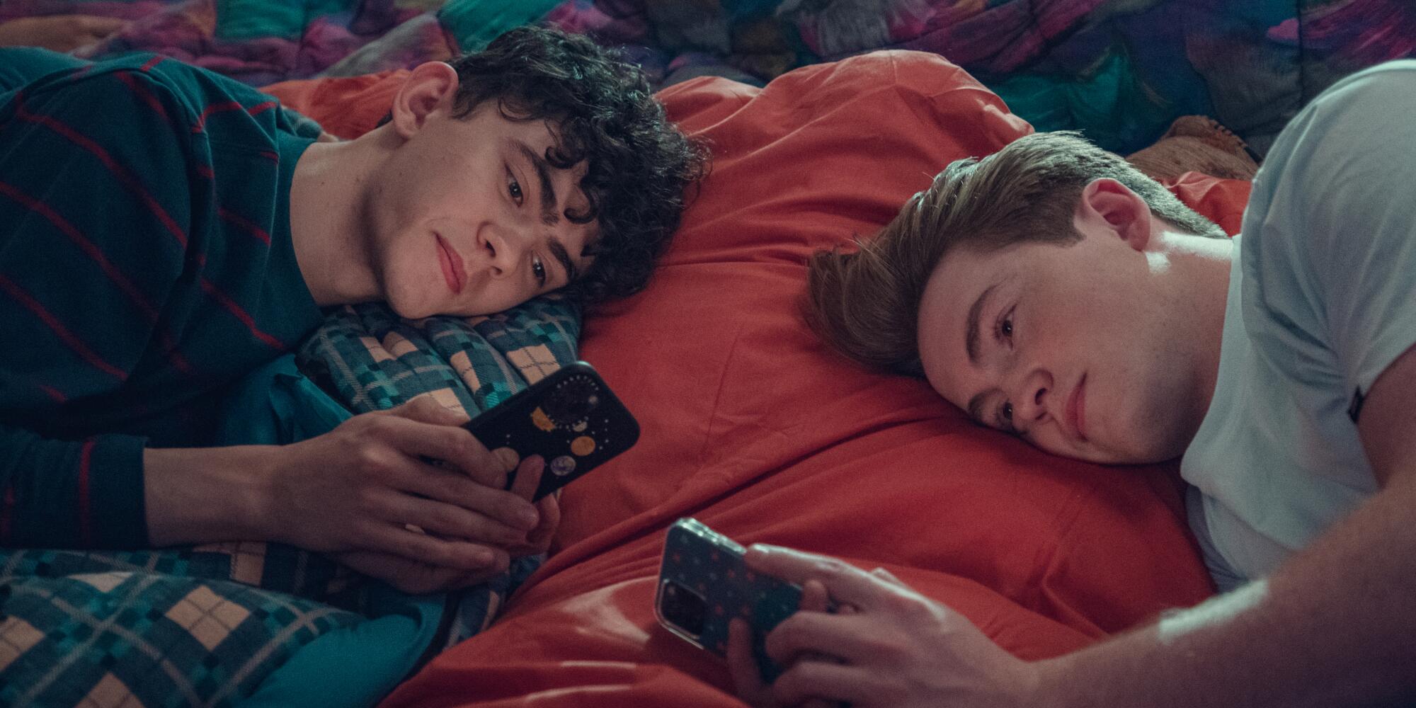 Two teen boys lie on sleeping bags near each other looking at their phones in "Heartstopper."