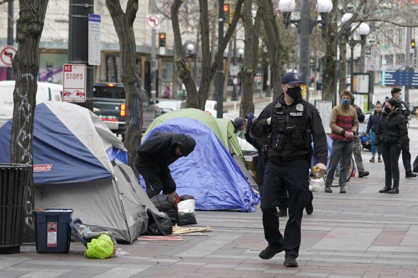 FILE - A Seattle police officer walks past tents used by people experiencing homelessness, March 11, 2022, during the clearing and removal an encampment in Westlake Park in downtown Seattle. The U.S. Justice Department and Seattle officials on Tuesday, March 28, 2023, asked a judge to end most federal oversight of the city's police department, saying its sustained, decade-long reform efforts are a model for cities around the country whose law enforcement agencies face federal civil rights investigations. (AP Photo/Ted S. Warren, File)
