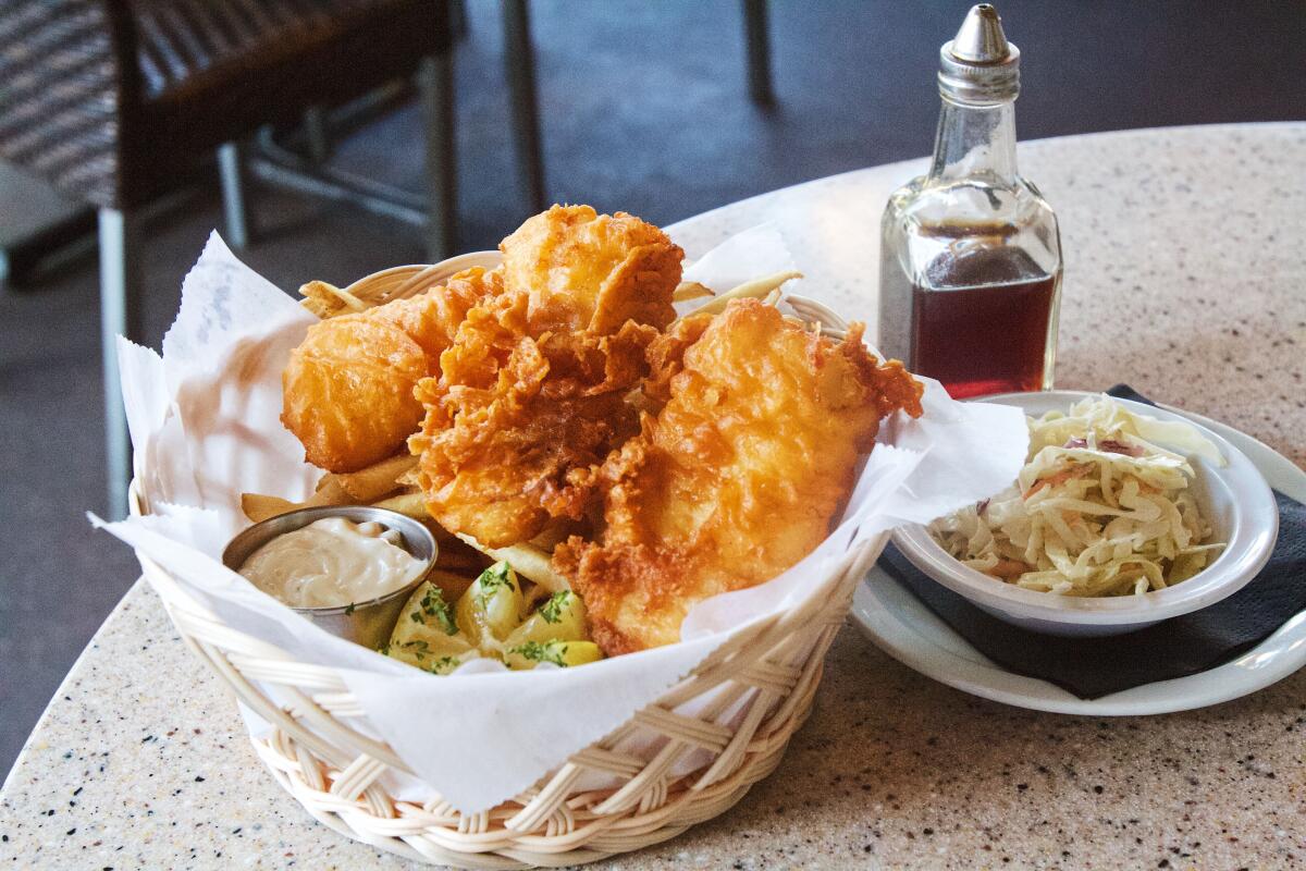 A basket of fish and chips on a tabletop. To the right are a bottle of vinegar and a side of slaw.