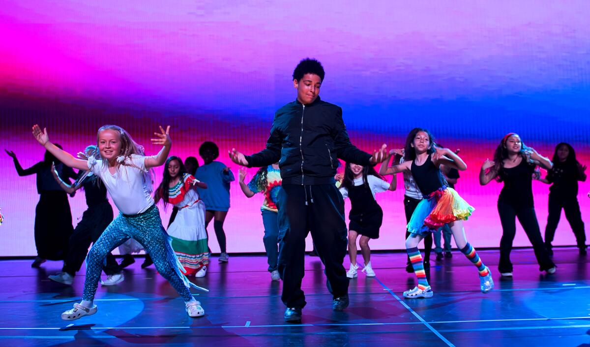 Children dance with arms raised on a stage with pink and blue lights.