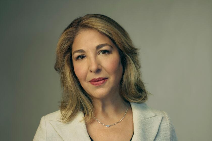 A woman with sandy blond hair stands in a white jacket over a black dress shirt.