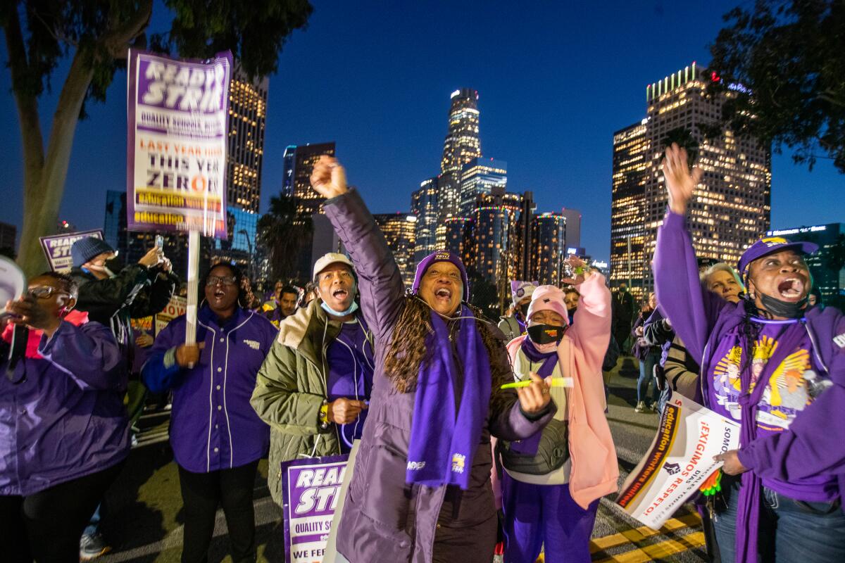 Demonstrators raise arms and hold signs with L.A. skyline in background.