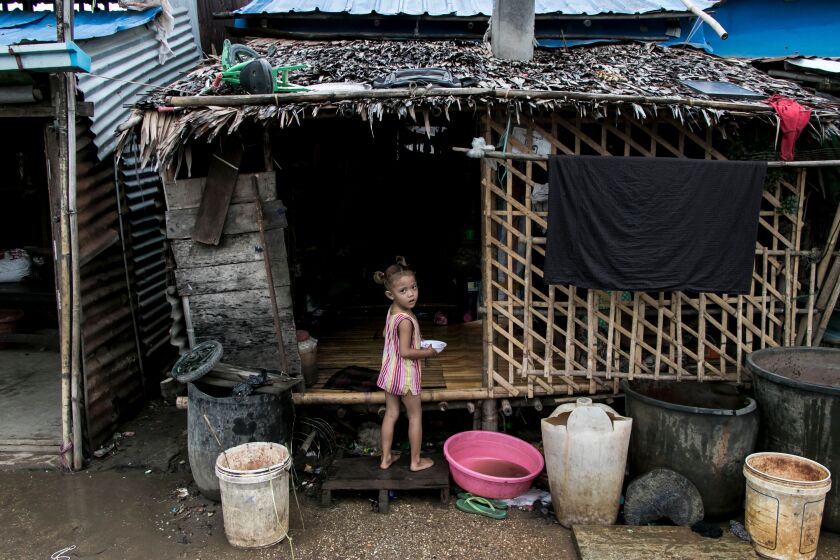 دختری در حومه یانگون در جلوی خانه خود ساخته شده از بامبو ، برگ و فلز موجدار.