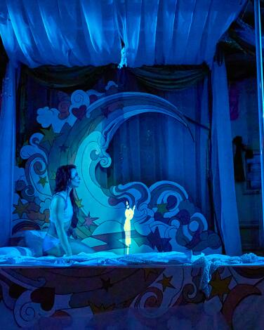 A woman on a bed with a projection of a small animated character. 