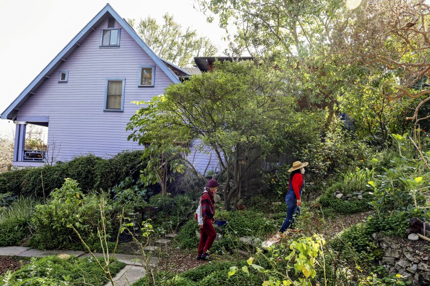 Two women walk on a greenery-lined path beside a house.