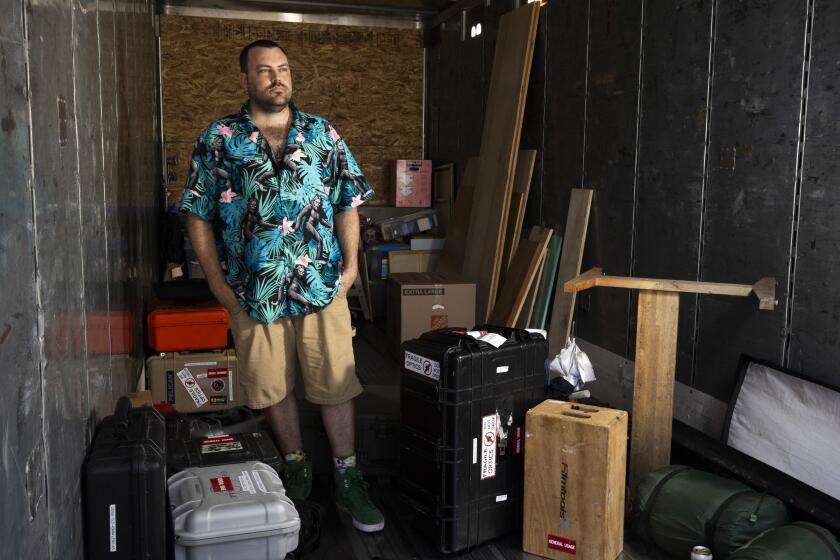 Photo of a man wearing a Hawaiian shirt in the back of a moving truck. He is surrounded by boxes.