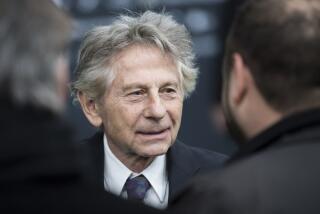 Roman Polanski wearing a black suit with a white undershirt and blue tie