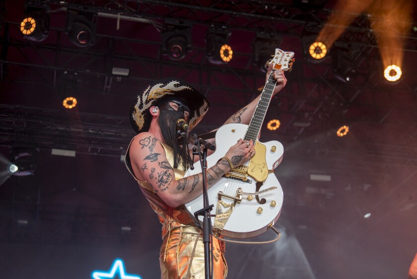 A man in a leather fringed mask and cowboy hat sings and plays guitar onstage