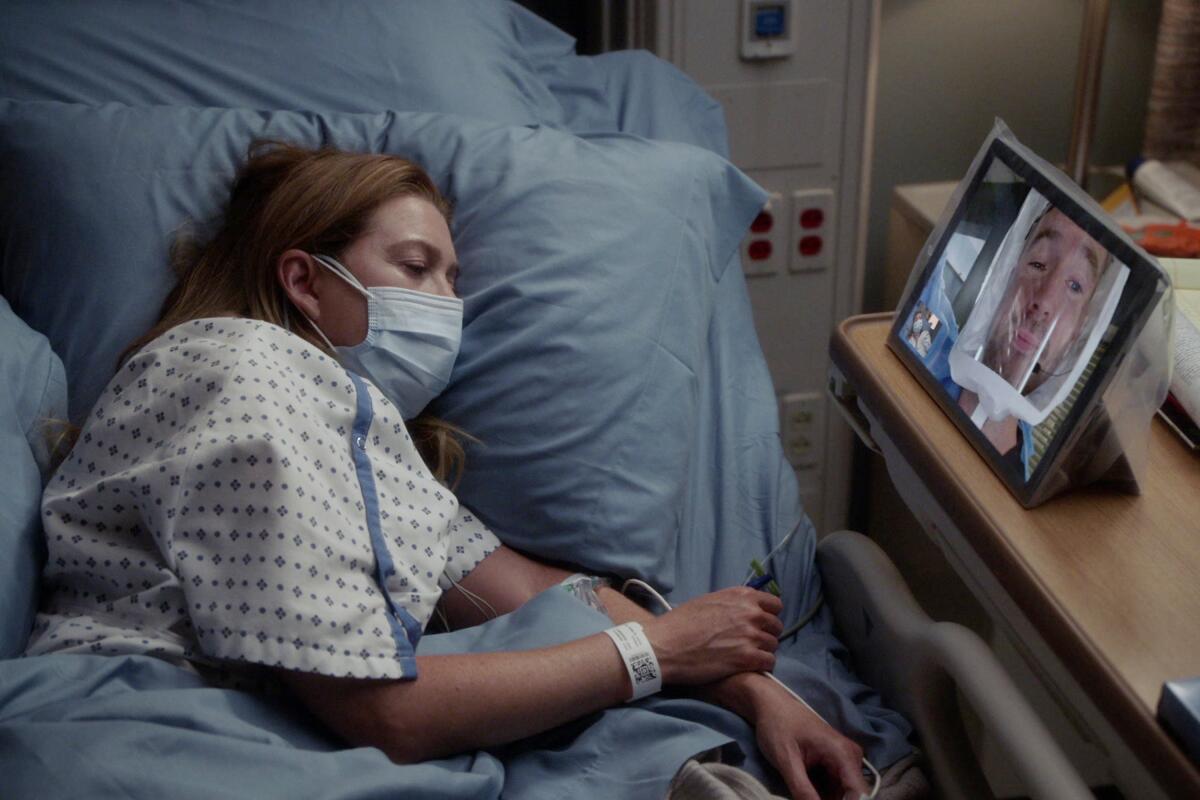 Ellen Pompeo as Meredith Grey in a mask lying in a hospital bed talking via video conference in a "Grey's Anatomy" episode.