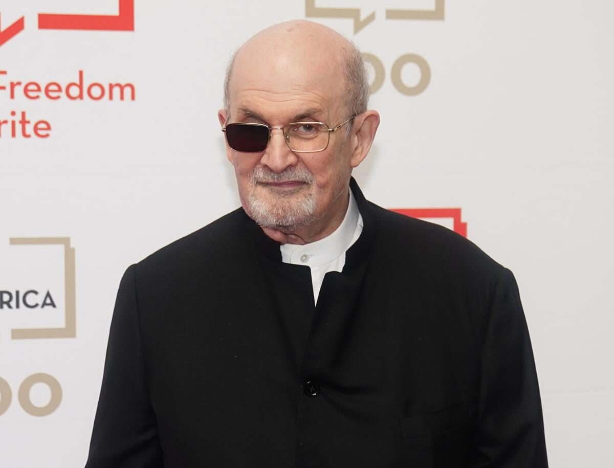 Salman Rushdie wears a black jacket with a white collared shirt under it as he poses for photos at a red carpet event