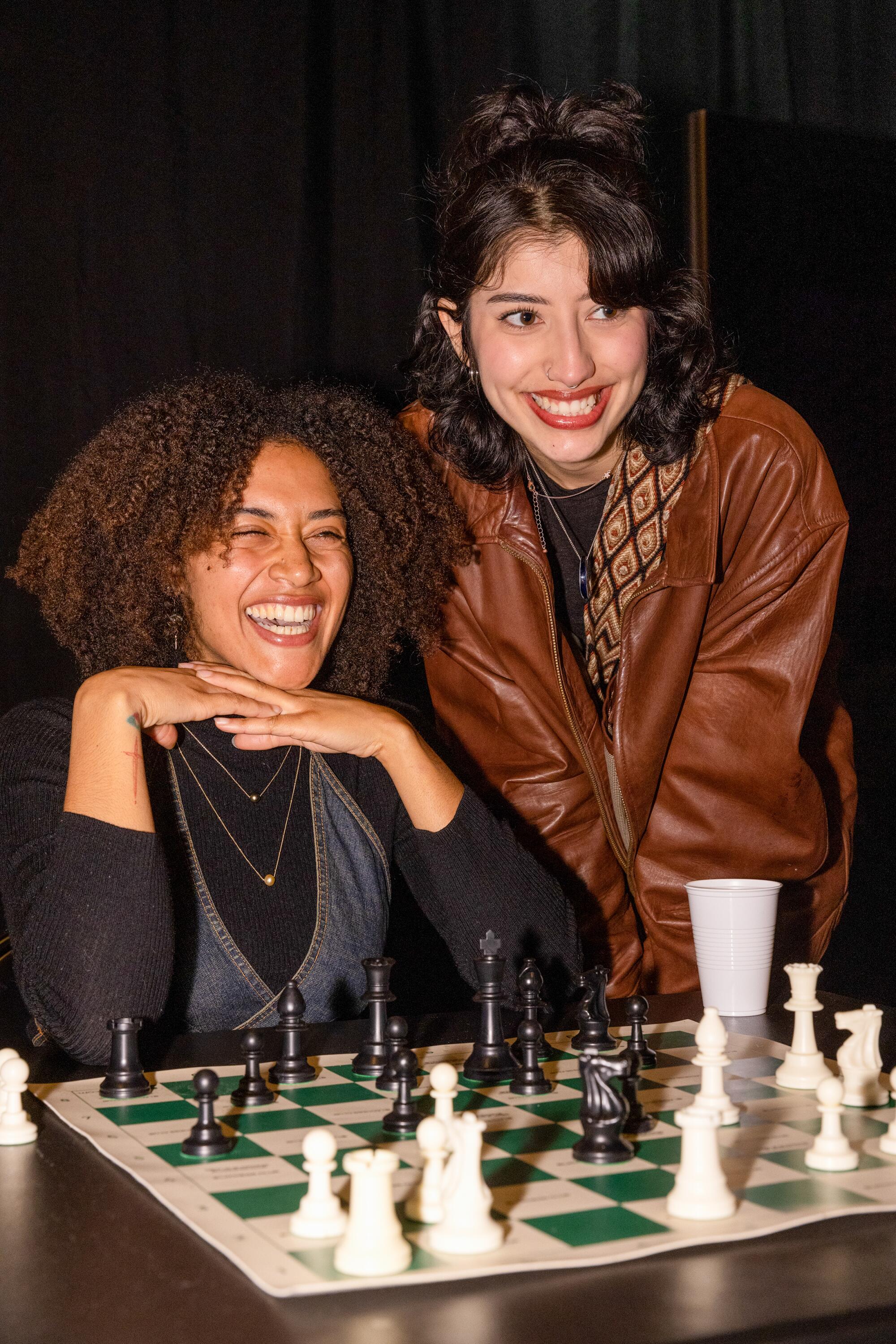 Two women, one seated, one standing, smile at the unseen person they're facing over a chess board