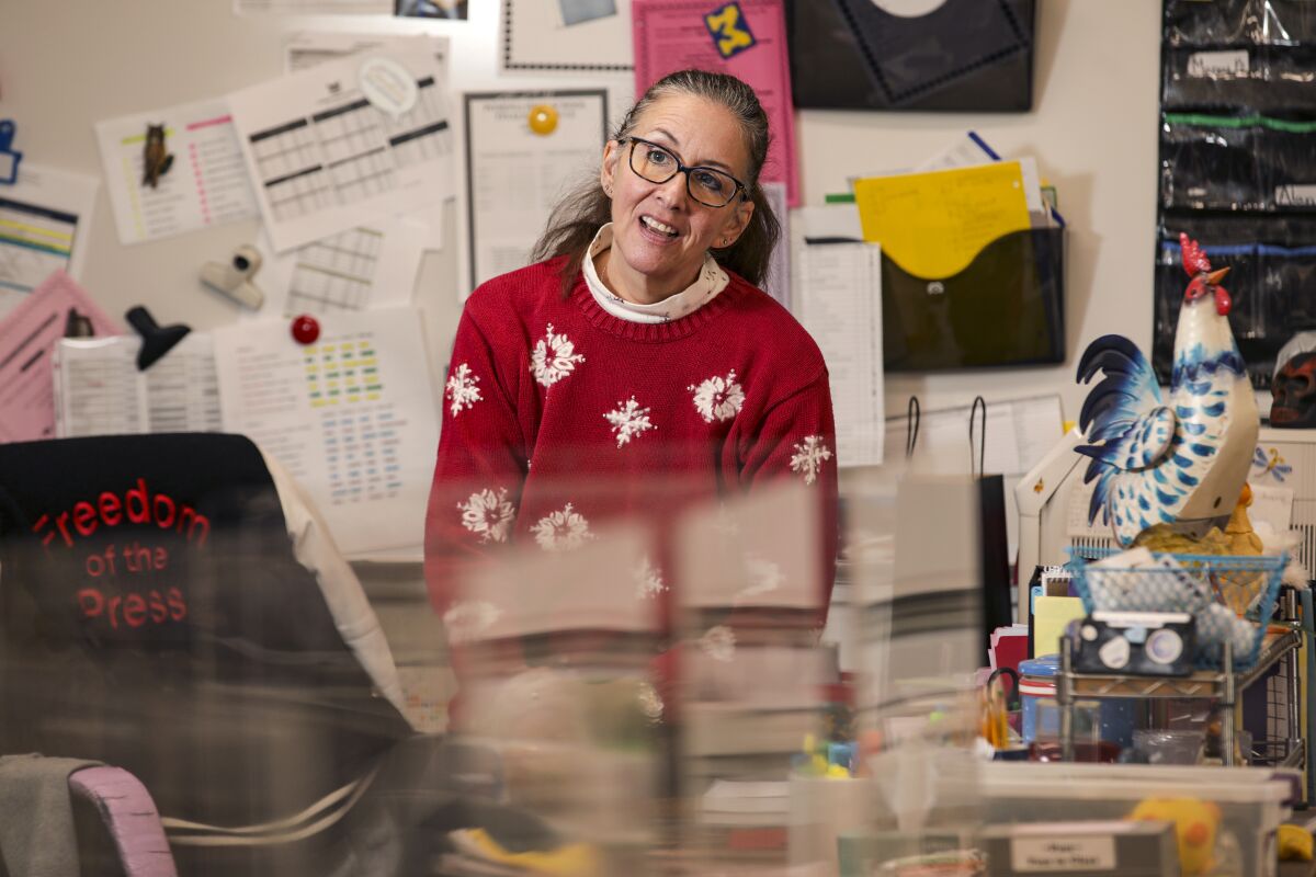 A woman in glasses and a red sweater with snowflakes sits at a desk in front of a bulletin board covered with paperwork.