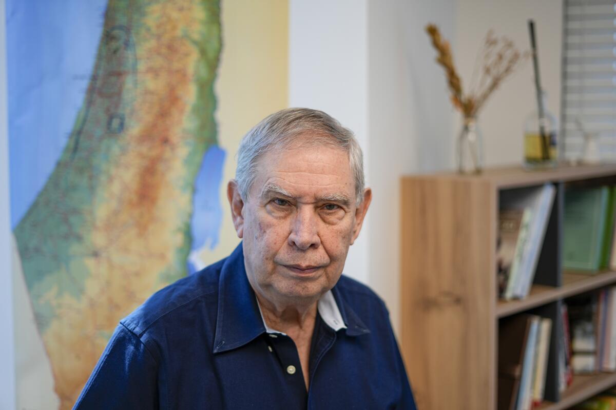 A man looks into the camera, in front of a large topographical wall map and a bookcase. 