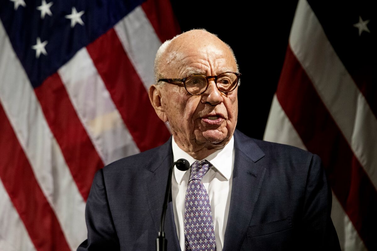 A man with glasses, in a tie and dark suit jacket, speaks at a microphone with U.S. flags behind him