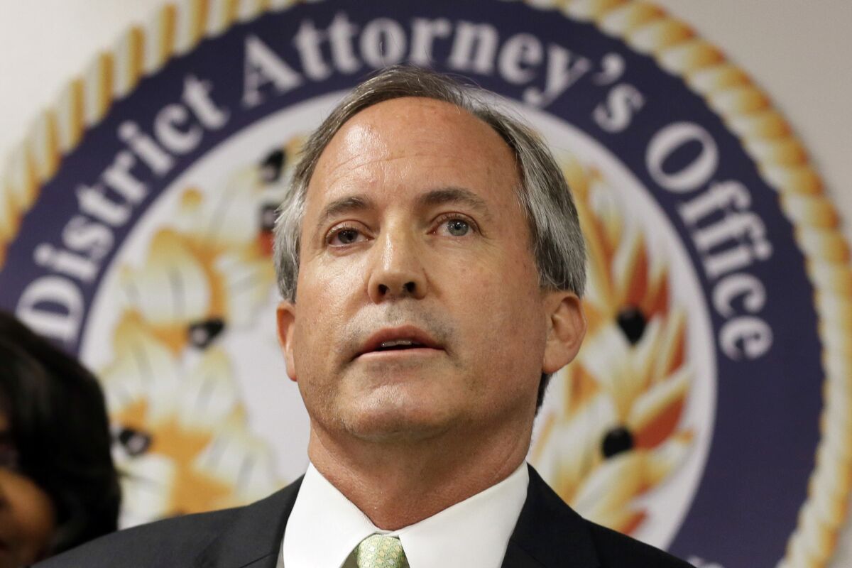 A closeup of Ken Paxton in front of a large state seal reading "District Attorney's Office"