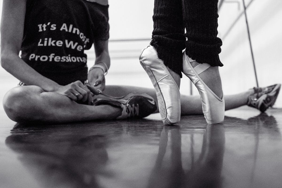 The feet of a dancer in pointe shoes next to a seated figure in a tee that reads, "It's Almost Like We're Professionals"