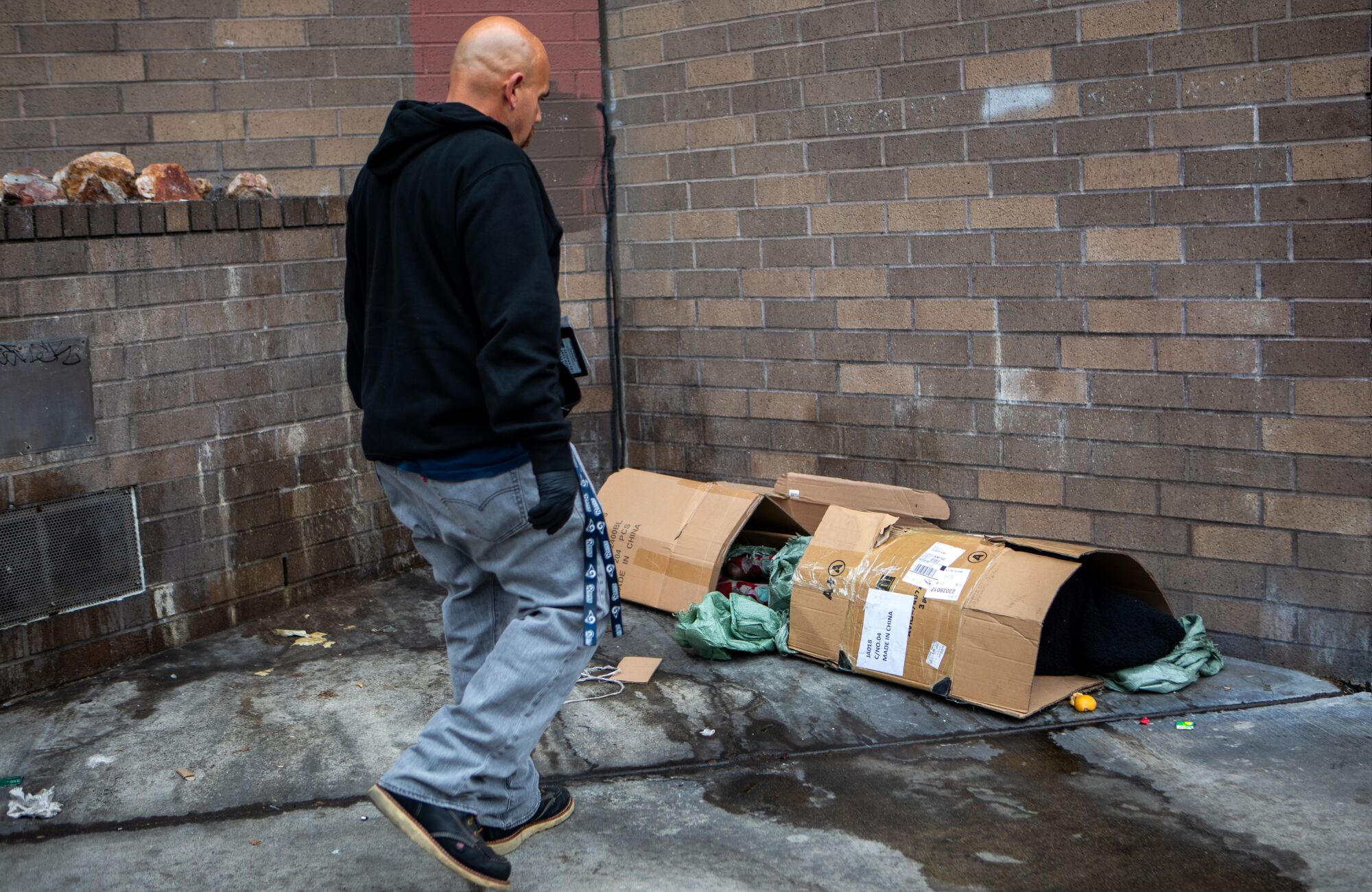 A man walks toward a person sleeping in a cardboard shelter.