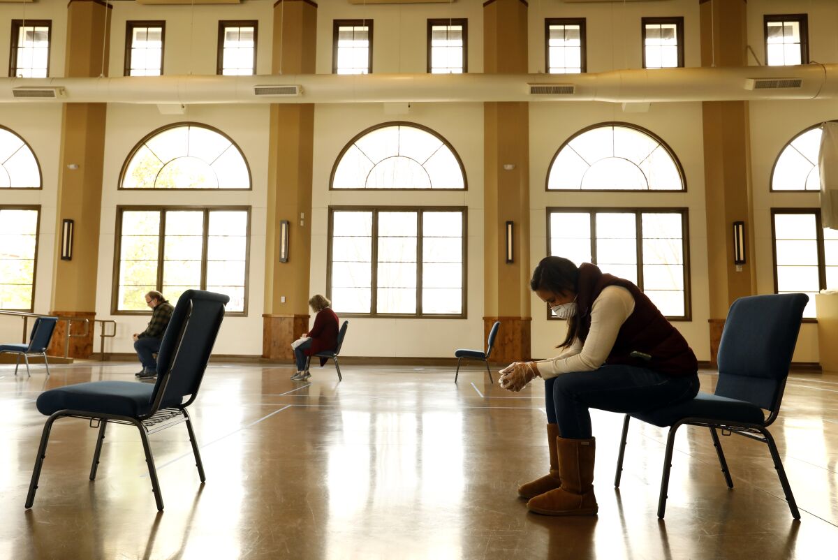 A woman wearing a mask sits in a chair and prays inside a building with other people seated on chairs in the background.