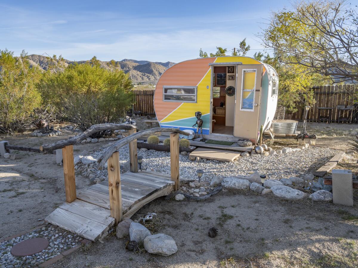 A teardrop trailer painted in rainbow colors sits at the end of a small foot bridge, desert foothills in the distance.