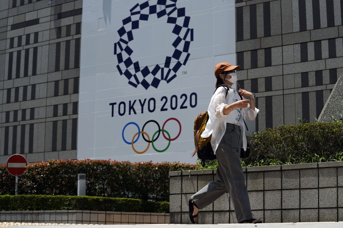 A woman wearing a protective mask and baseball cap walks past a Tokyo Olympics sign.