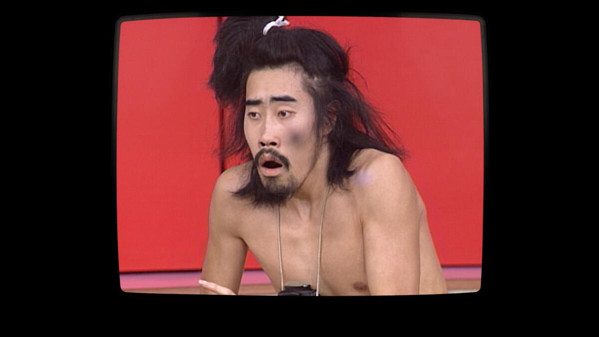A shirtless man on a TV set, squatting before a red backdrop.
