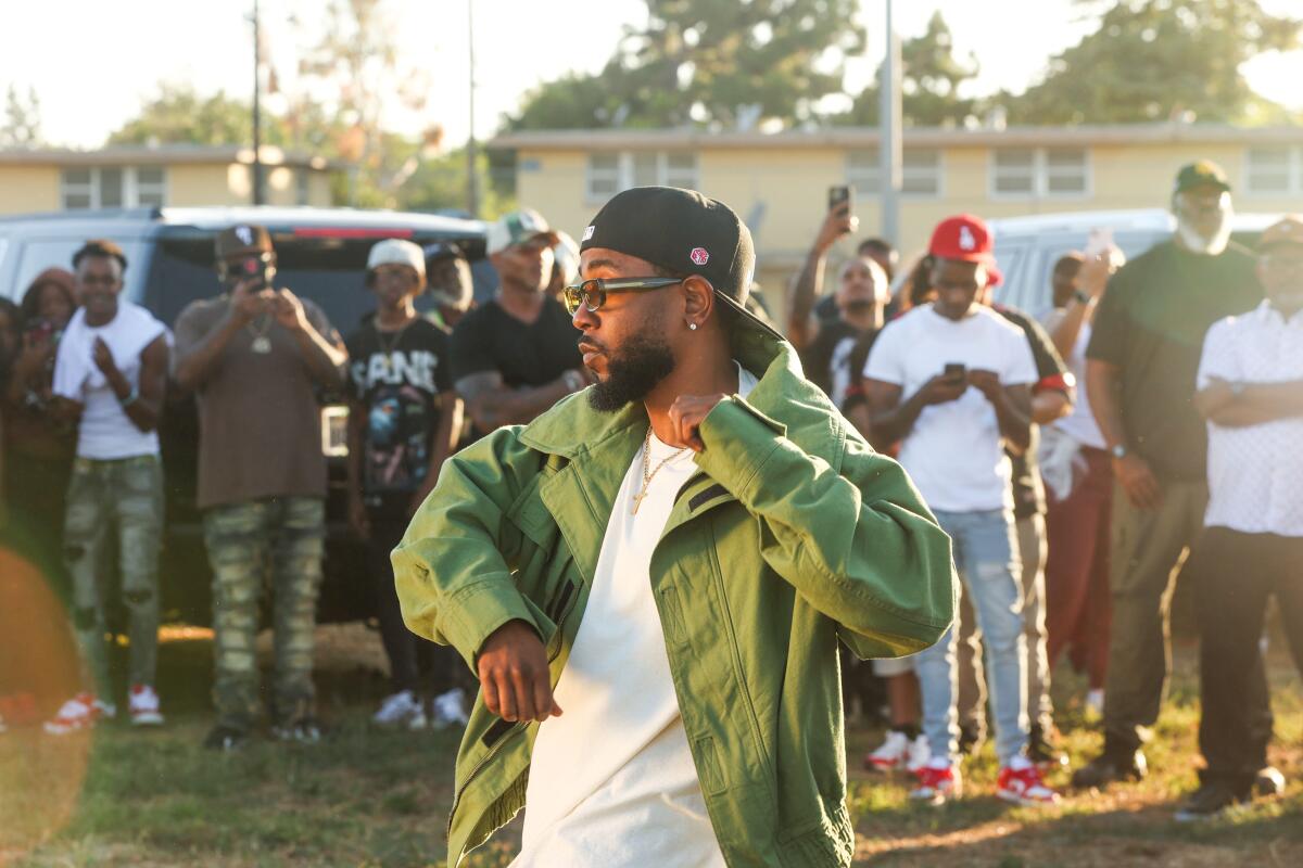 Kendrick Lamar in a green jacket, hat and white T-shirt dancing outdoors in front of a group of people