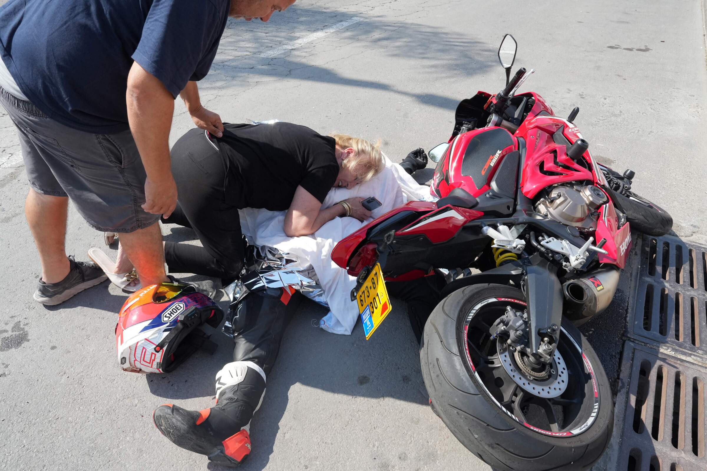 A man stands over a woman holding onto a body lying on the ground, covered by a sheet, next to a red motorcycle 
