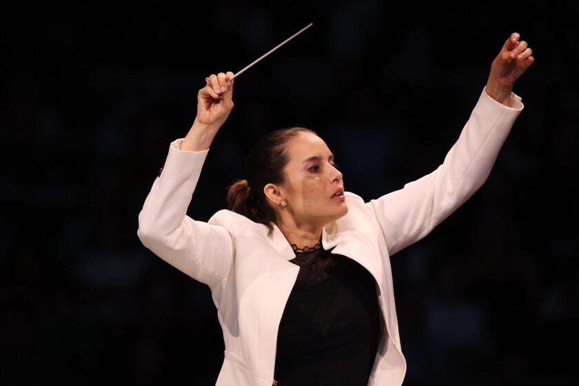 A woman wearing a long white jacket is seen in close-up leading an orchestra with a baton.