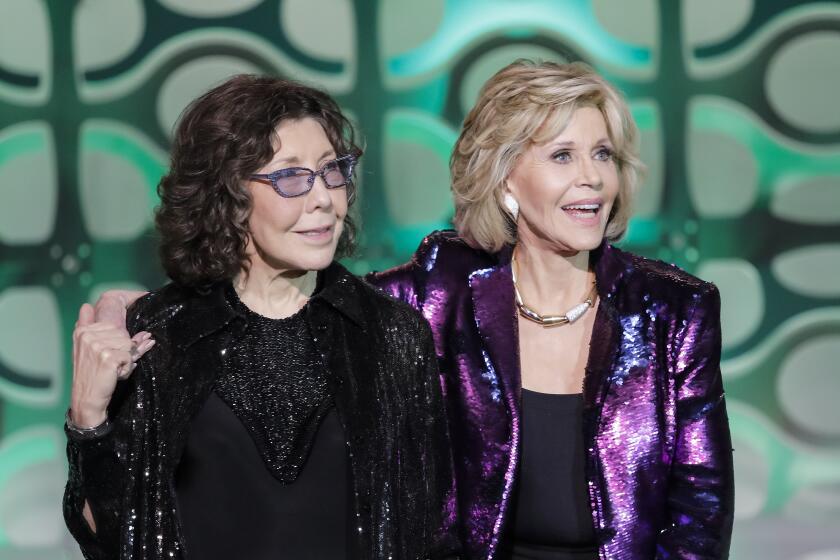A mature woman stands on stage with her arm around the shoulders of another mature woman wearing glasses