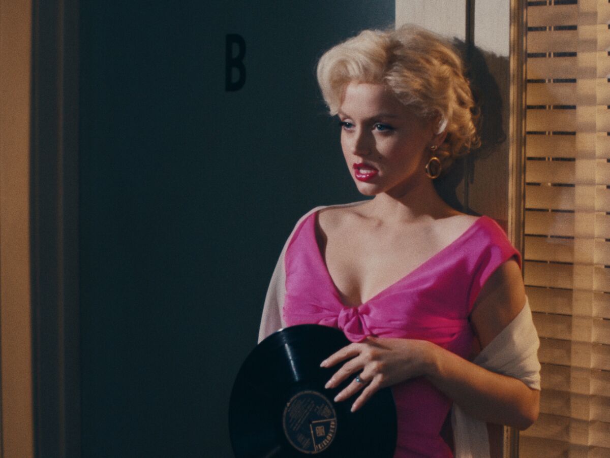 A woman with short, platinum blonde hair wears a pink dress and holds a vinyl record in one scene "Blond."