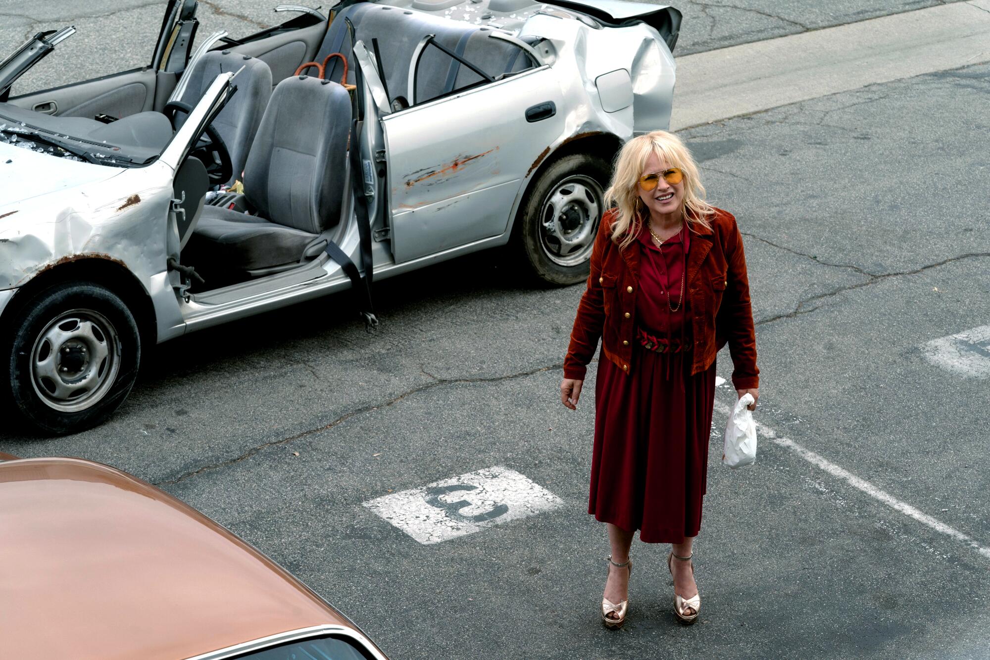A blonde woman in a maroon jacket and dress stands in front of a beat up silver car with no roof.