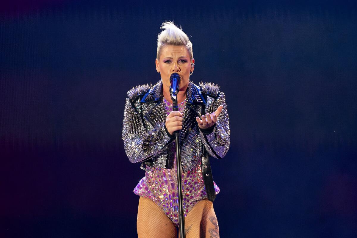 Pink standing behind a mic and talking, wearing a bedazzled pink bodysuit and sparkly black jacket with fuzzy shoulders