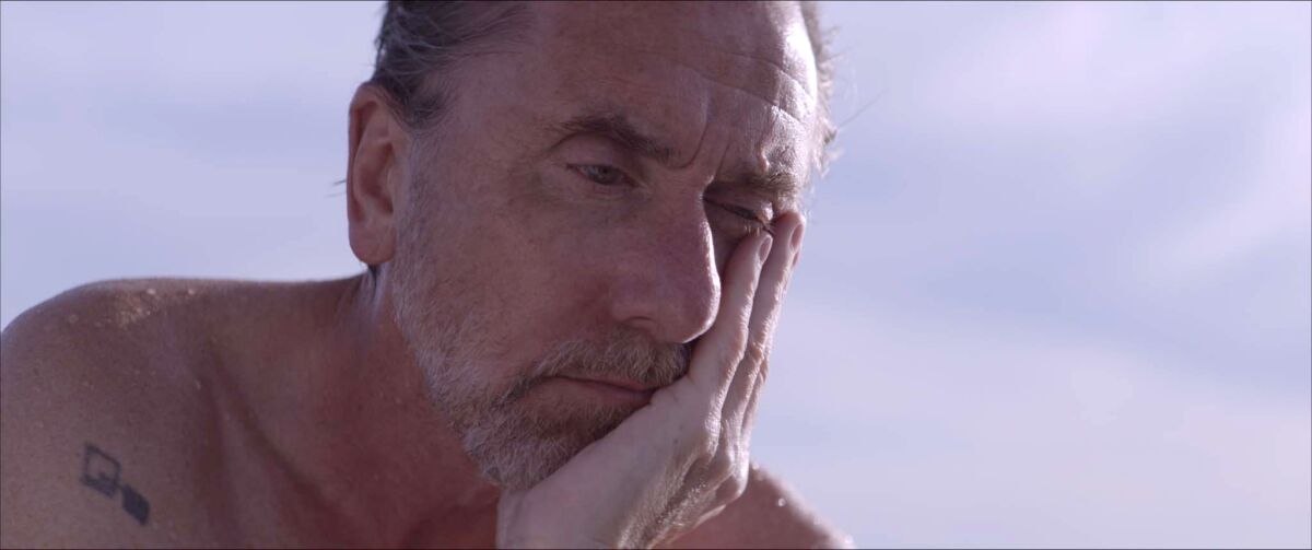A shirtless man sits in the sun and leans his chin into one hand.