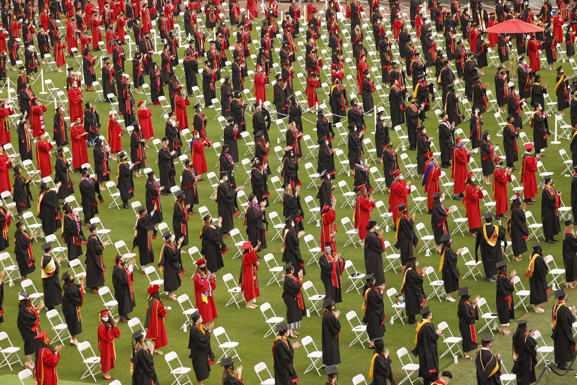 Dozens of students in red or black gowns stand and clap in front of spaced-out white folding chairs on a field