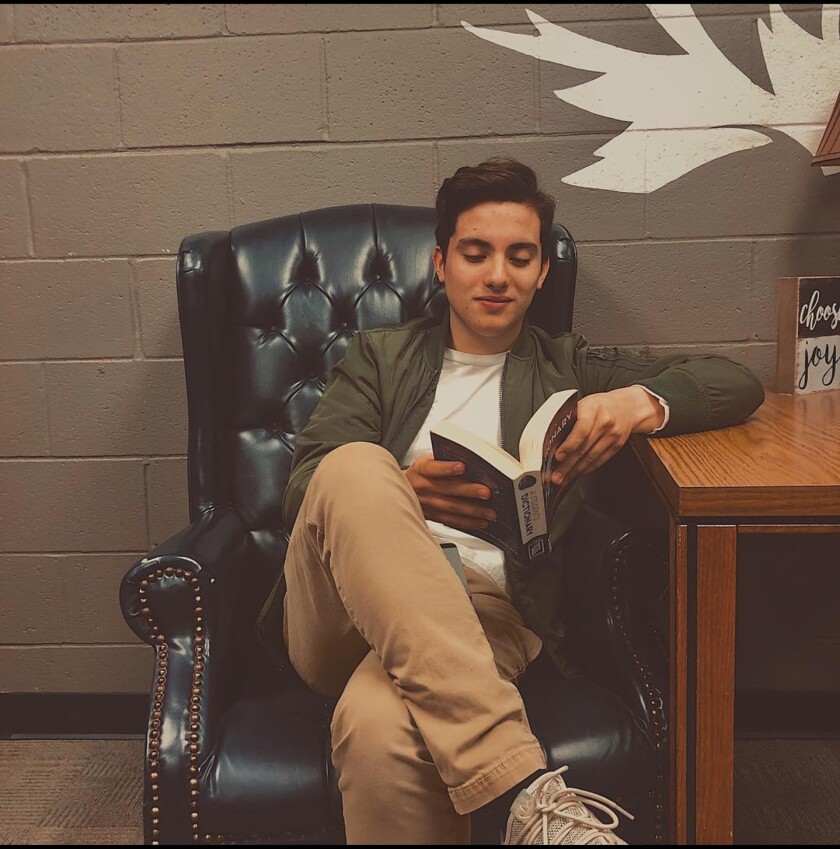 A young man sits in a black armchair while reading a book.