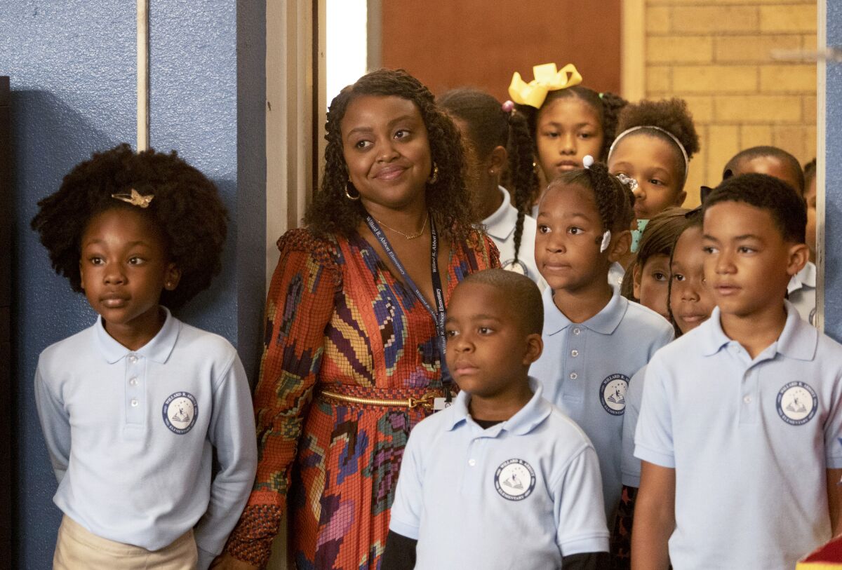 A woman in a patterned dress stands among a group of elementary students wearing school uniforms.