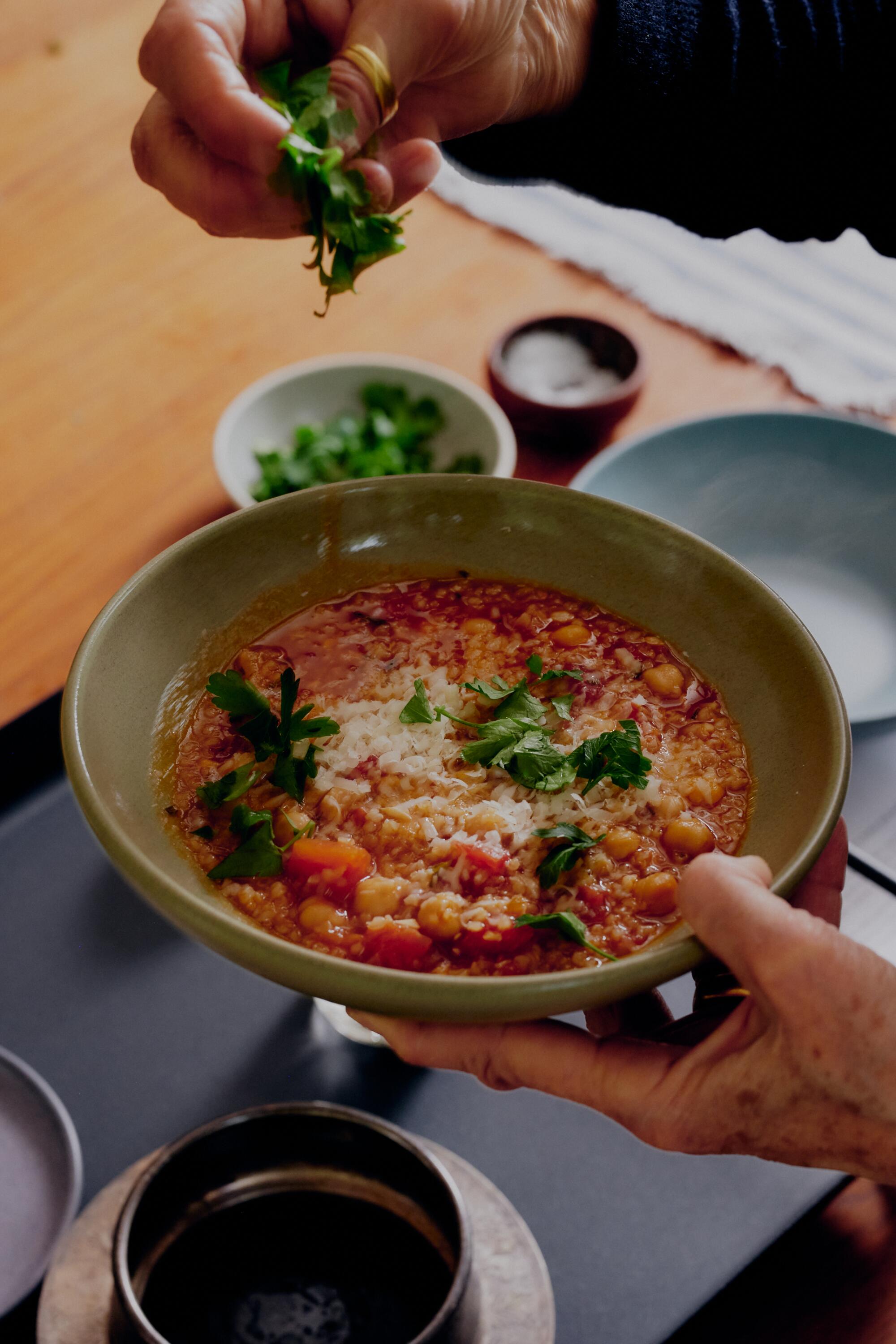 Hands garnishing a bowl of Roman-style Chickpea and Tomato Soup With Bulgur
