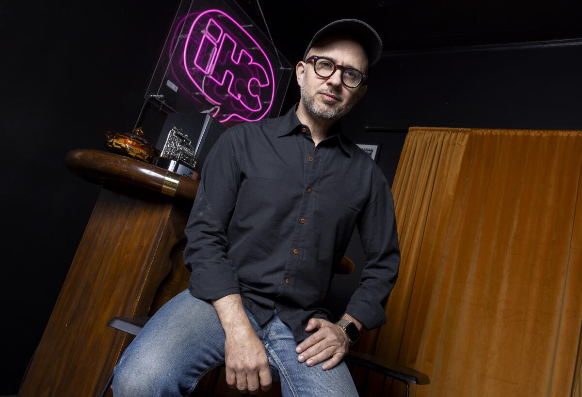 A man in blue jeans, black shirt, glasses and black ballcap sits in front of a neon word balloon with letters I H C